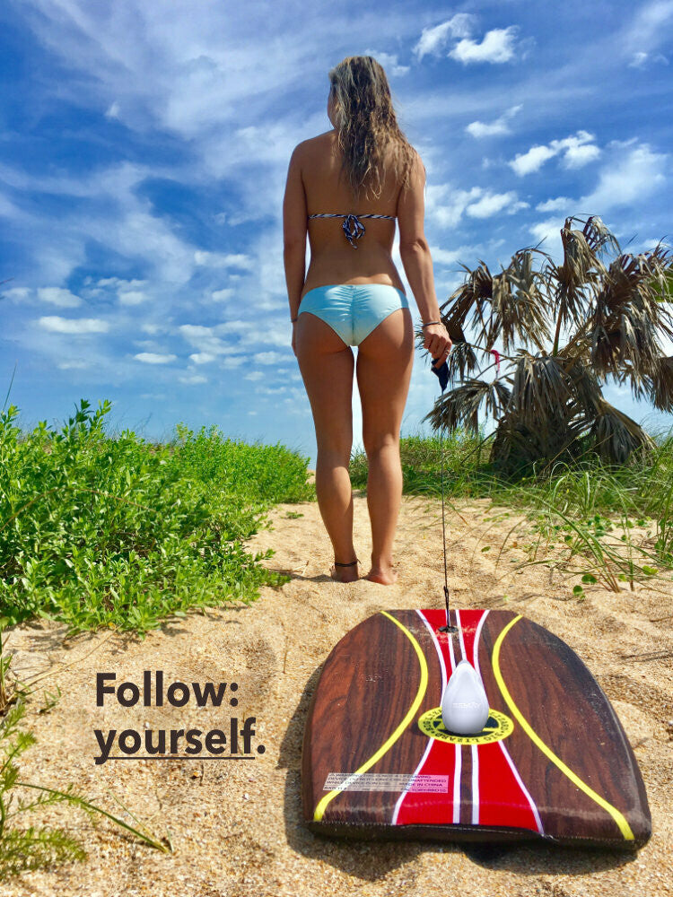 Woman in a bikini at the beach pulling a serf board across sand with green plants and blue skies with a white REMAY shave gel bar on the board in the foreground.