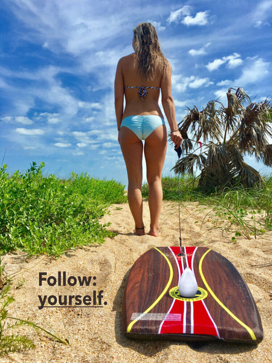 Woman in a bikini at the beach pulling a serf board across sand with green plants and blue skies with a white REMAY shave gel bar on the board in the foreground.